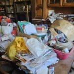 Salon encombré avec piles de papiers et objets divers.
