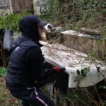 Personne soulève objet dans jardin envahi par la végétation.